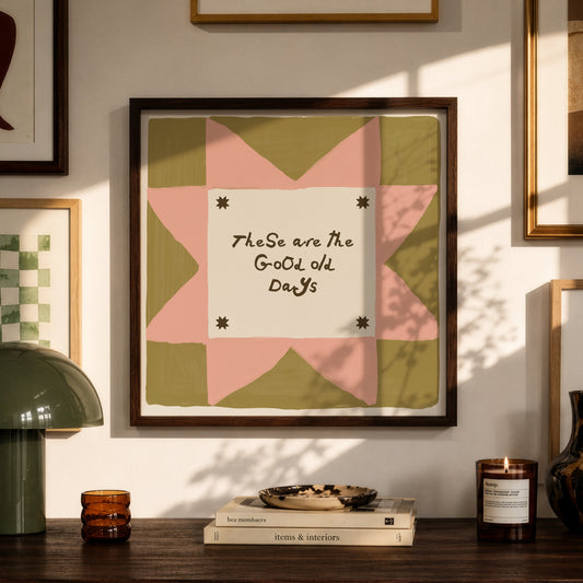 Framed wall art with a quilt-style geometric design in pink, olive green, and cream, featuring the handwritten text 'TheSe are the Good Old Days.' The artwork is displayed in a styled room setting above a wooden console table with a green lamp, books, and candles.
