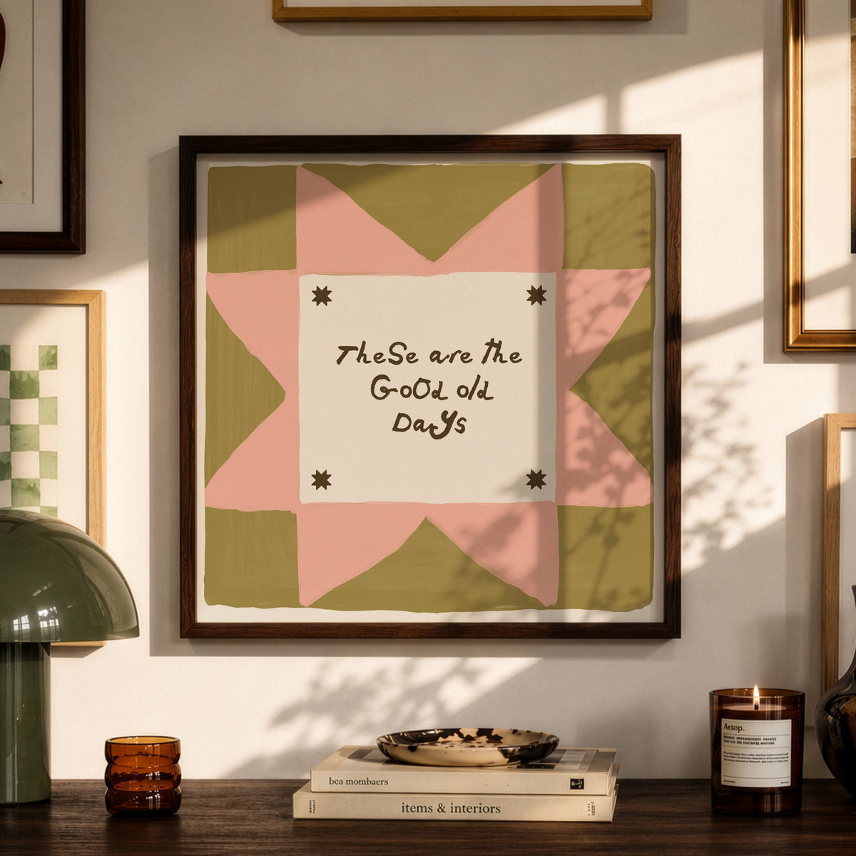 Framed wall art with a quilt-style geometric design in pink, olive green, and cream, featuring the handwritten text 'TheSe are the Good Old Days.' The artwork is displayed in a styled room setting above a wooden console table with a green lamp, books, and candles.