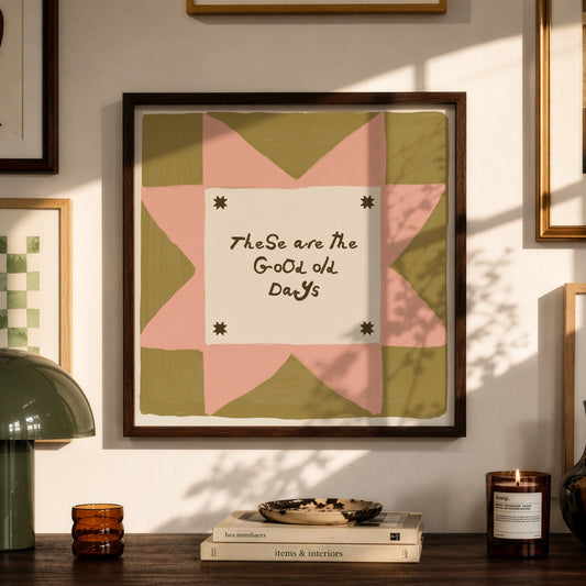 Framed wall art with a quilt-style geometric design in pink, olive green, and cream, featuring the handwritten text 'TheSe are the Good Old Days.' The artwork is displayed in a styled room setting above a wooden console table with a green lamp, books, and candles.