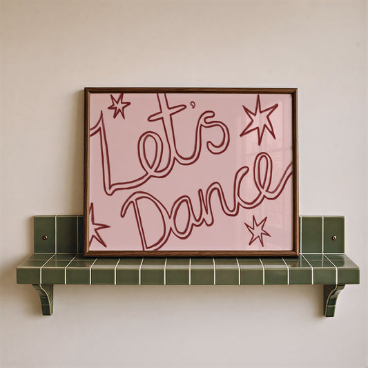 A framed wall art print with the words “Let’s Dance” in red script on a light pink background, leaning against a beige wall on a green tiled shelf.