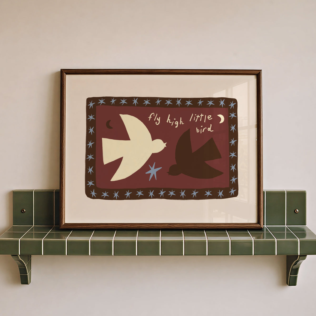 A framed art print featuring a stylized cream and dark brown bird illustration with the words 'fly high little bird' on a burgundy background, displayed on a green tiled shelf against a light beige wall.