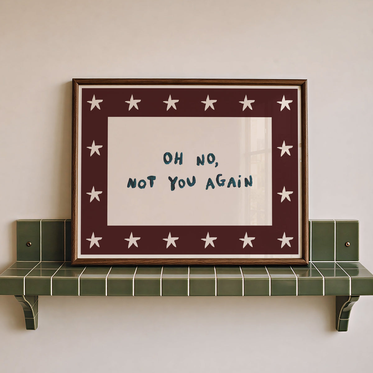 Framed wall art with the handwritten phrase “OH NO, NOT YOU AGAIN” in dark blue on a white center, surrounded by a dark red border with white stars, displayed on a green tiled shelf against a light beige wall.