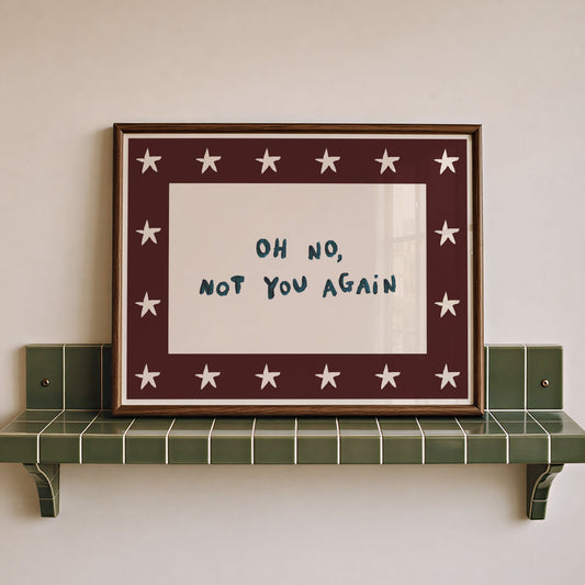 Framed wall art with the handwritten phrase “OH NO, NOT YOU AGAIN” in dark blue on a white center, surrounded by a dark red border with white stars, displayed on a green tiled shelf against a light beige wall.