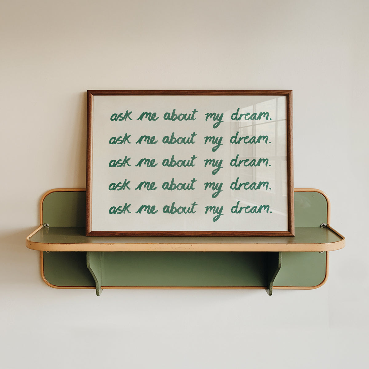 A framed typographic wall art print with the phrase 'ask me about my dream.' repeated in green script on a white background, displayed on a green floating shelf against a light beige wall.