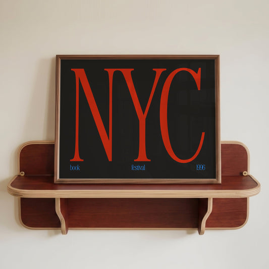 A framed art print featuring the word 'NYC' in large red serif letters on a black background, displayed on a wooden floating shelf against a plain off-white wall.