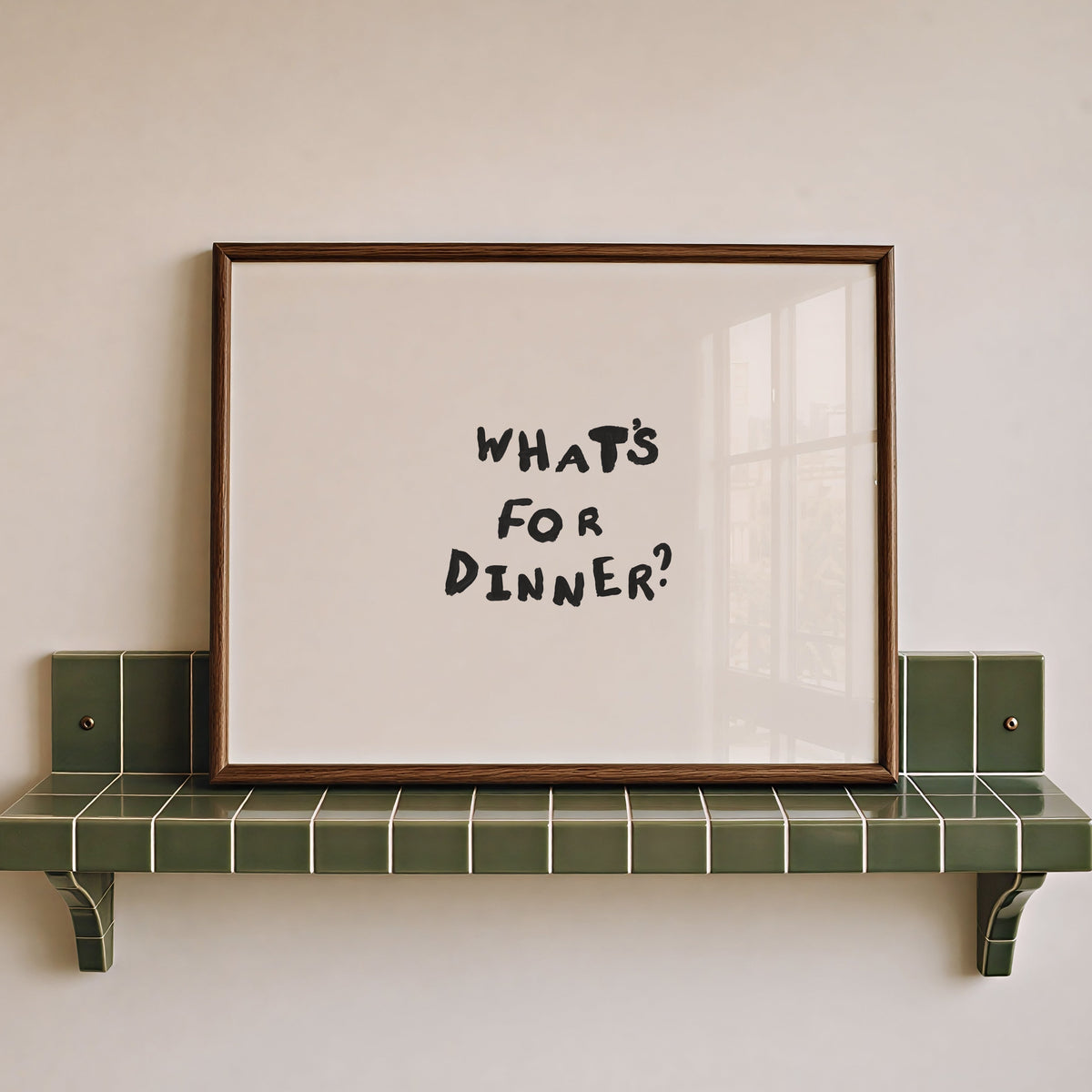 A framed typographic wall art print reading “WHAT’S FOR DINNER?” in black handwritten-style lettering on an off-white background, leaning against a beige wall on a green tiled shelf.